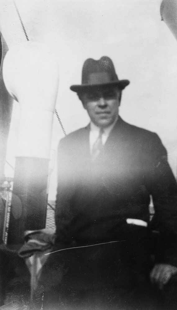 Blurry, overexposed black and white photograph due to light leakage. It depicts a man leaning in a nautical setting. He is dressed in a neat suit and a black fedora.