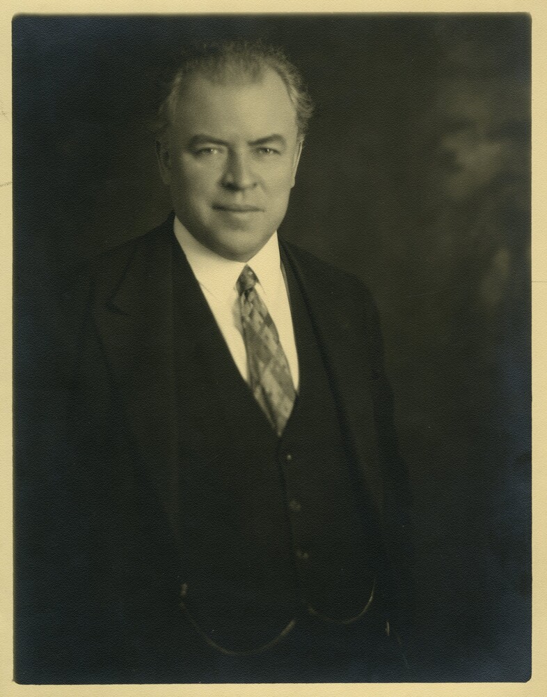 Black and white photograph depicting a man facing forward. He is wearing a three-piece suit.