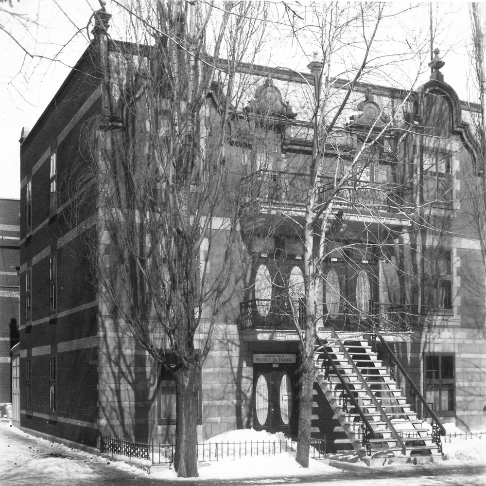 Black and white photograph depicting the facade of a large triplex partially hidden behind trees. The ground is covered with snow. The eight-unit building also has two front balconies.