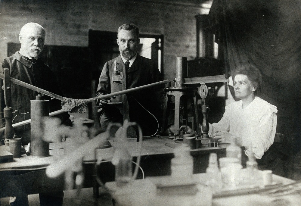 Black and white photograph of a laboratory with several scientific instruments on a table. Three people are seated at the table. Two men are standing facing the camera on the left and a woman is seated manipulating laboratory instruments on the left.