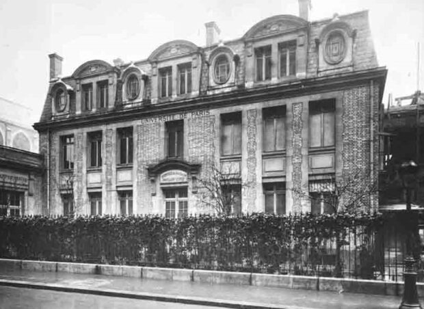 Black and white photograph of the facade of a three-story brick building with large dormer windows on the third floor. Above the entrance, it reads Université de Paris. The building is surrounded by a metal fence through which a vine grows.