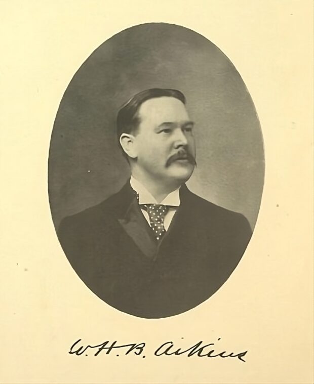 Black and white photograph of a mustachioed man wearing a suit and tie. The photograph is bordered by an oval shape and the man's signed name in cursive letters is inscribed below: W. H. B. Aikins.