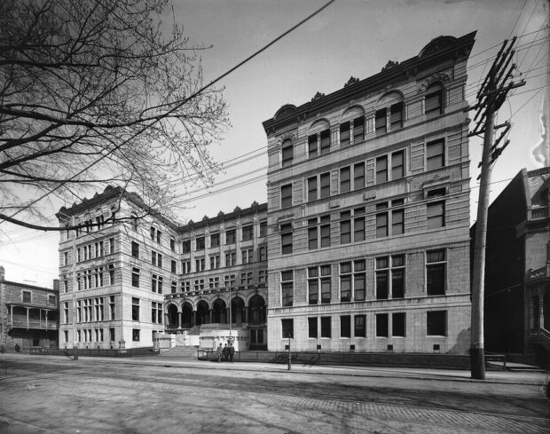 Black and white photograph of a five-story imposing building built like a hook with its interior facing the street. In the center, on the sidewalk opposite the building, three men are standing and looking at the camera.