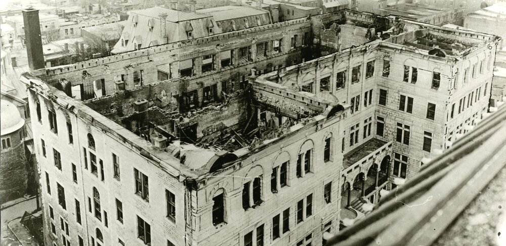 Black and white photograph, taken from above, of a large five-story building, the tallest of which was completely destroyed following a fire.