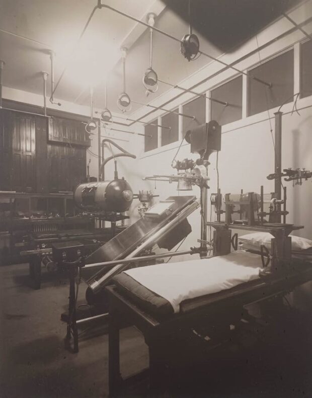 Black and white photograph depicting a room filled with equipment and tables set up as hospital beds. The devices are varied, some resembling barrels, others old photographic cameras.