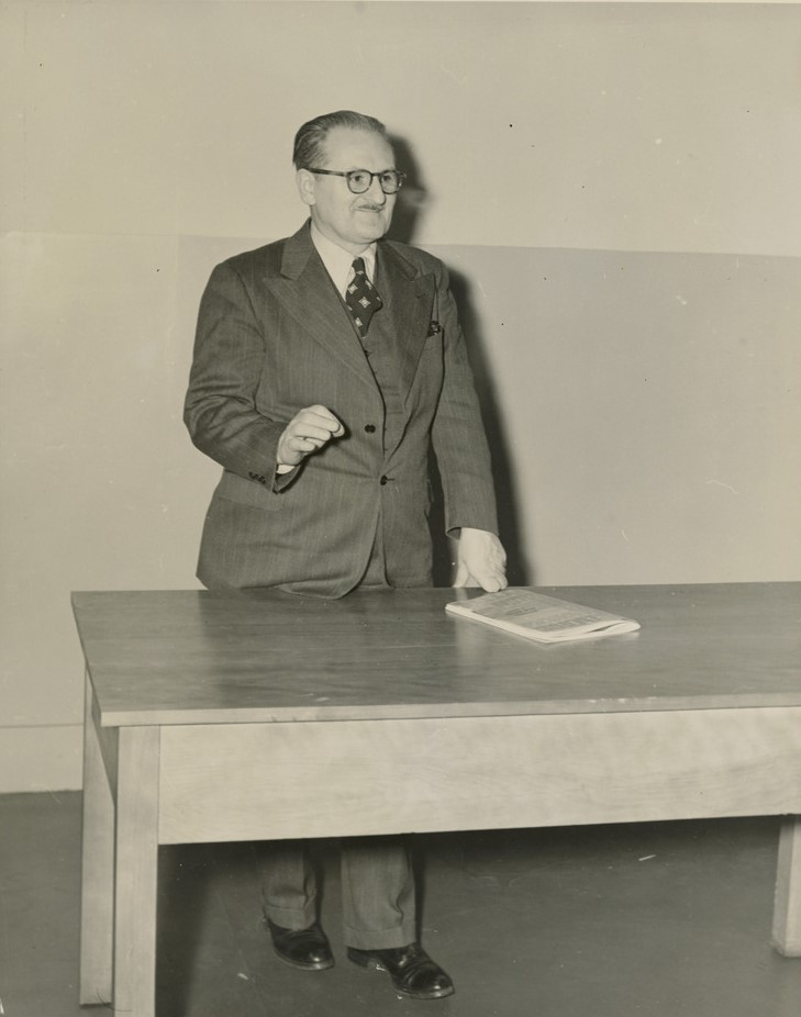 Black and white photograph depicting a man standing behind a wooden table wearing a three-piece suit. The man wears glasses and seems to be gesturing something. A random magazine is the only item placed on the table.