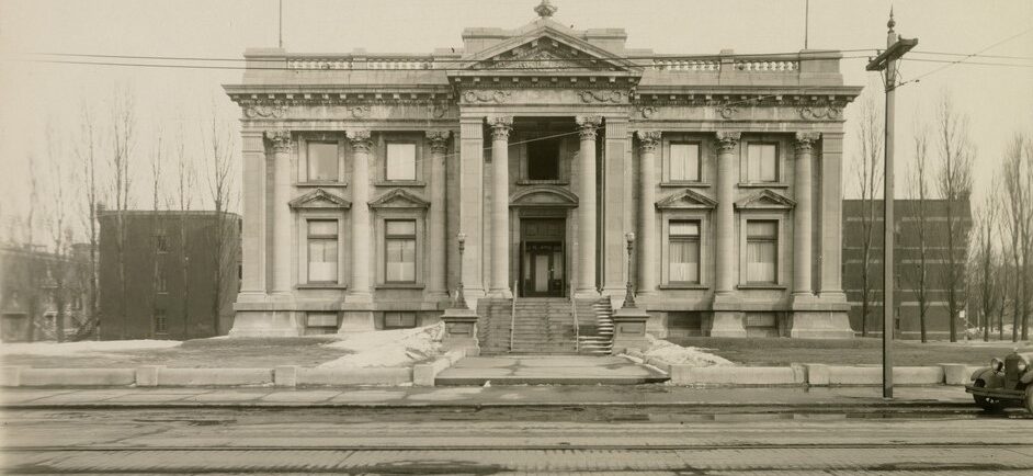 Black and white photograph of an imposing Beaux-Arts style building with a concrete path leading to the stairs and central doors. In the foreground is the brick-paved street.