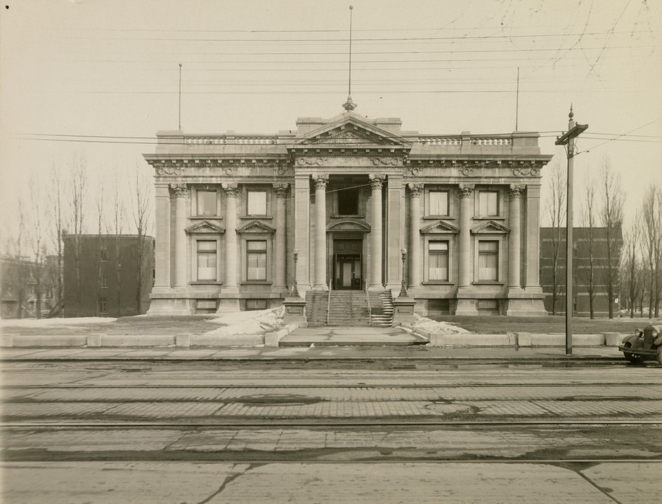 Black and white photograph of an imposing Beaux-Arts style building with a concrete path leading to the stairs and central doors. In the foreground is the brick-paved street.