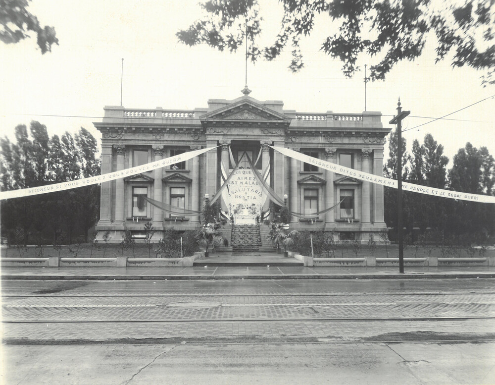 Black and white photograph of a large Beaux-Arts style building. Its facade is decorated with banners attached to its columns. The banners read: Lord, if you will, you can heal me. Lord, just say the word and I will be healed. The one you love is sick. Salutaris hostia.