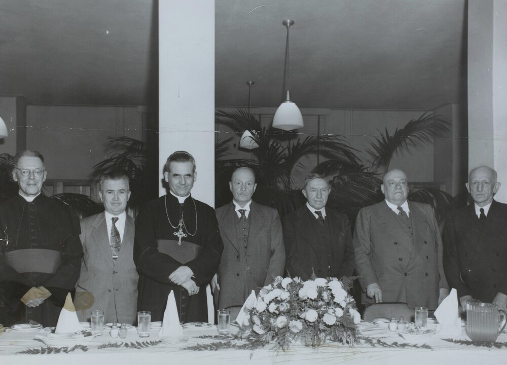 Black and white photograph depicting seven men standing in a large hall behind a banquet table. Most of them are dressed in three-piece suits, but two of them are wearing cassocks.