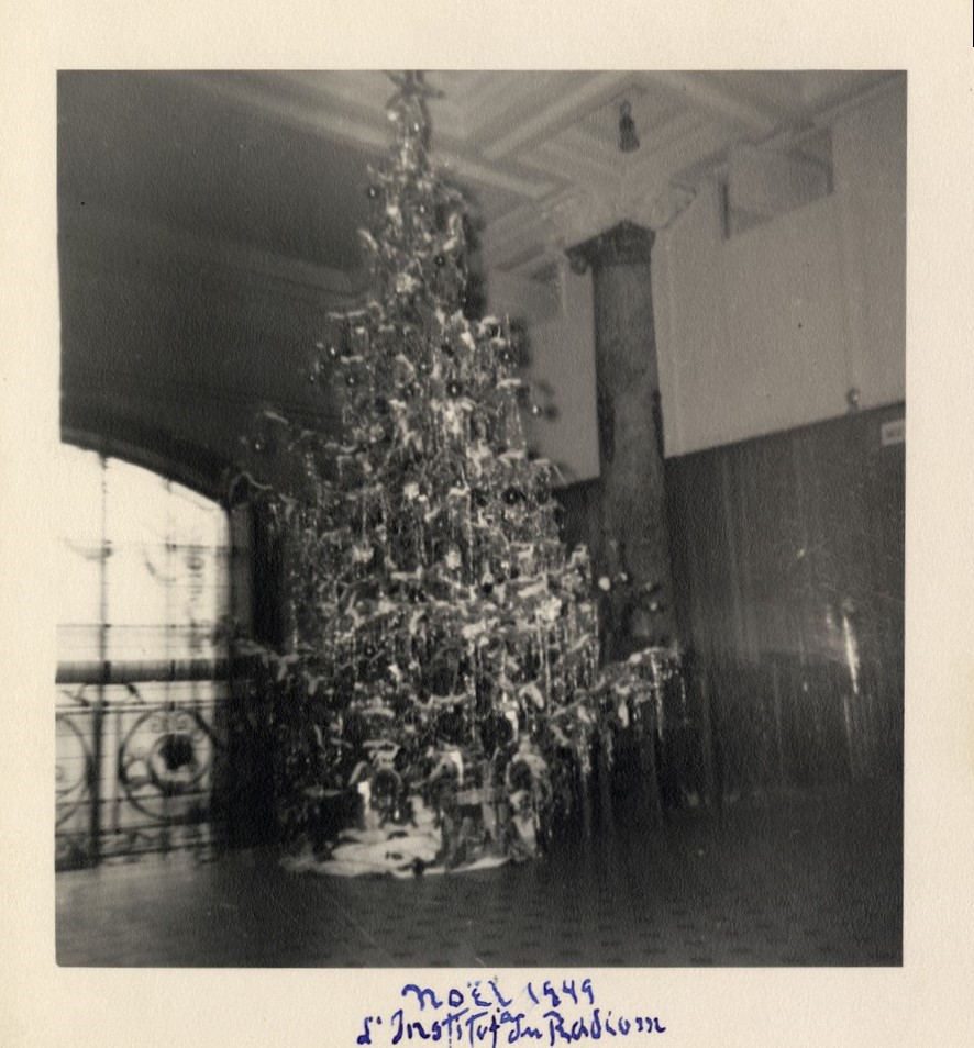 Black and white photograph, slightly blurry, of a richly decorated Christmas tree located in a hall.