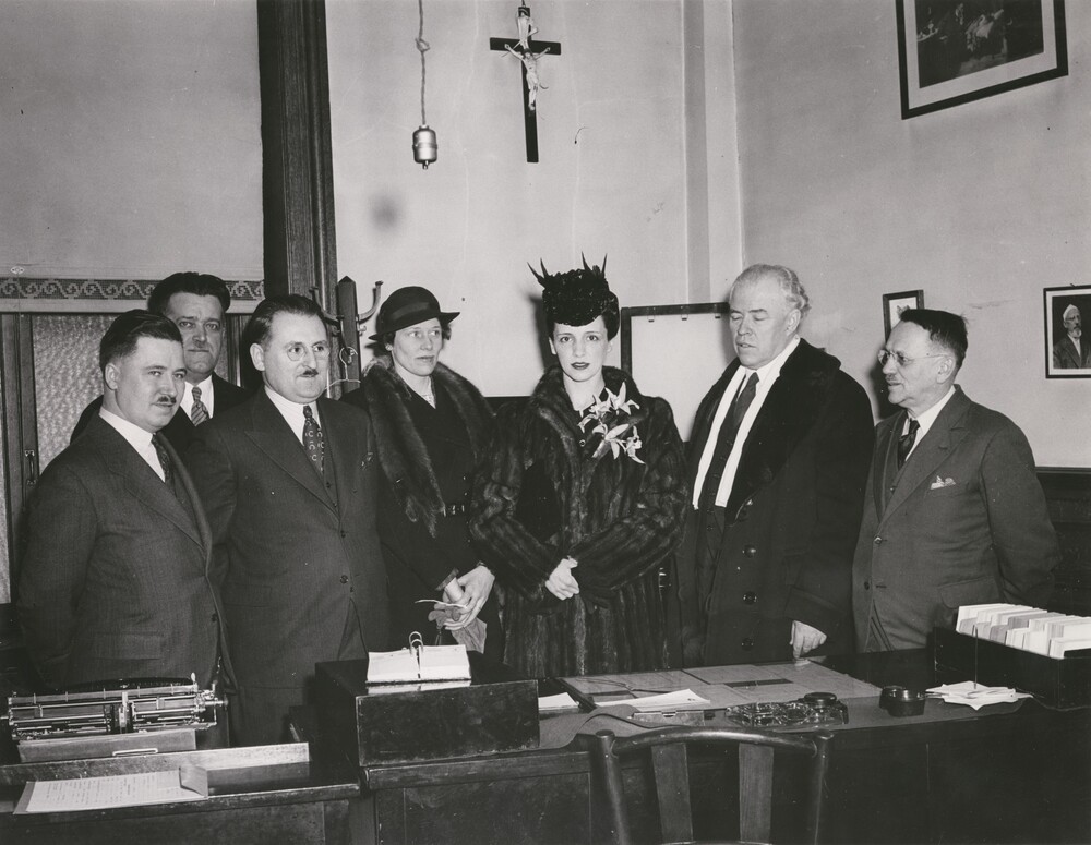 Black and white photograph depicting seven well-dressed people behind a desk. There are five men and two women in the center. The group is in a high-ceilinged room with a window, photos, and a crucifix on the walls. The only furniture is a desk and a wooden chair.