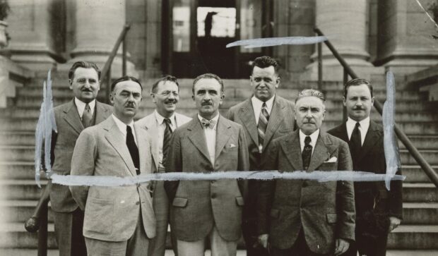 Black and white photograph depicting seven men in suits in frowomennt of stone stairs in the background leading to the main door of the brick and stone building, of which only the base is visible.