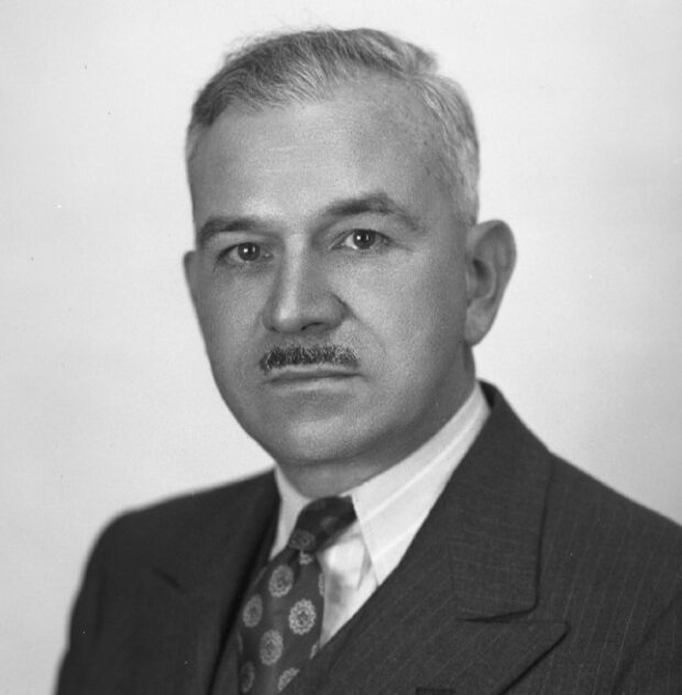 Black and white photograph depicting a man facing forward in a suit and tie. He has short graying hair and a small mustache.