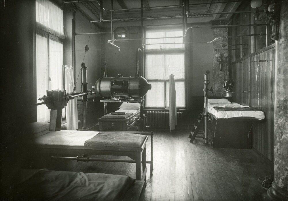 Black and white photograph of a room with a high ceiling well lit by two large windows filtered by thin curtains. Three beds are clearly visible in the image and arranged to accommodate machine installations on top. The three machines look like metal boxes and one of them looks like a large barrel lying lengthwise.