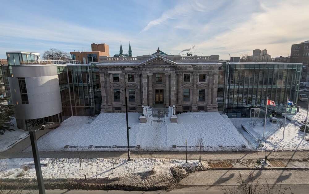 Color photograph depicting a building with a facade made of large gray stones accompanied by Beaux-Arts-inspired pillars. Modern extensions have been added along its sides, with facades made of glass and metal sheets.