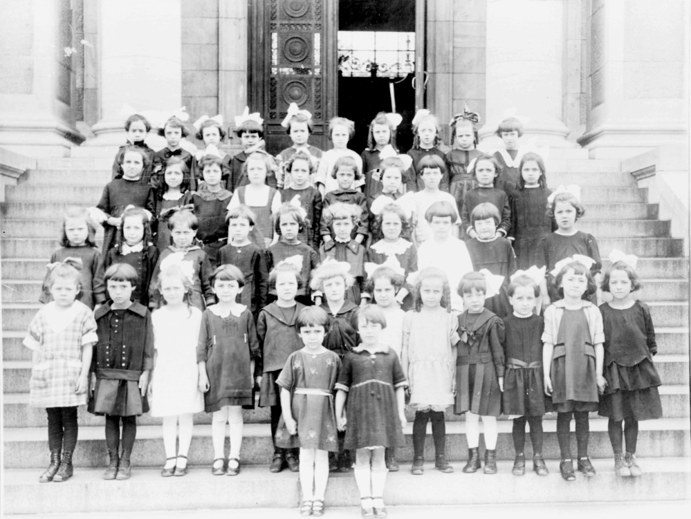 Black and white photograph depicting about forty girls arranged in four rows on the stone steps leading to the main door of the stone building.