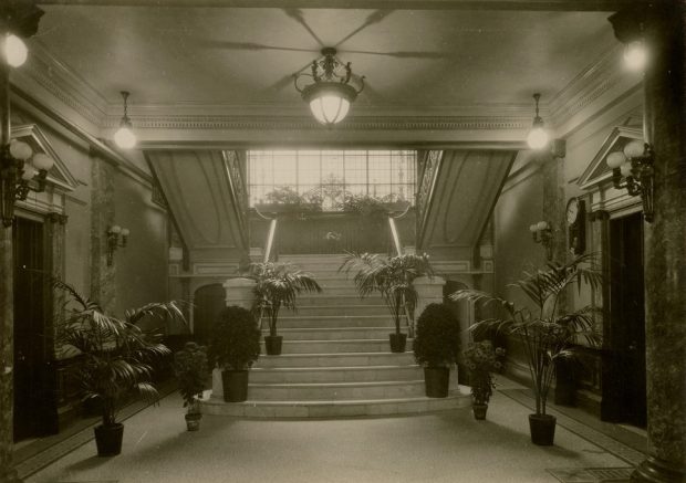 Black and white photograph of an entrance hall leading to a staircase. The hall has Beaux-Arts features with marble pillars and white walls. Eight potted plants are arranged to form a path to the stairs.