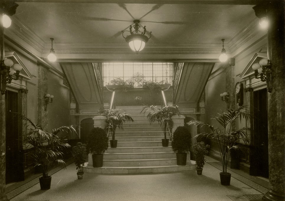 Black and white photograph of an entrance hall leading to a staircase. The hall has Beaux-Arts features with marble pillars and white walls. Eight potted plants are arranged to form a path to the stairs.