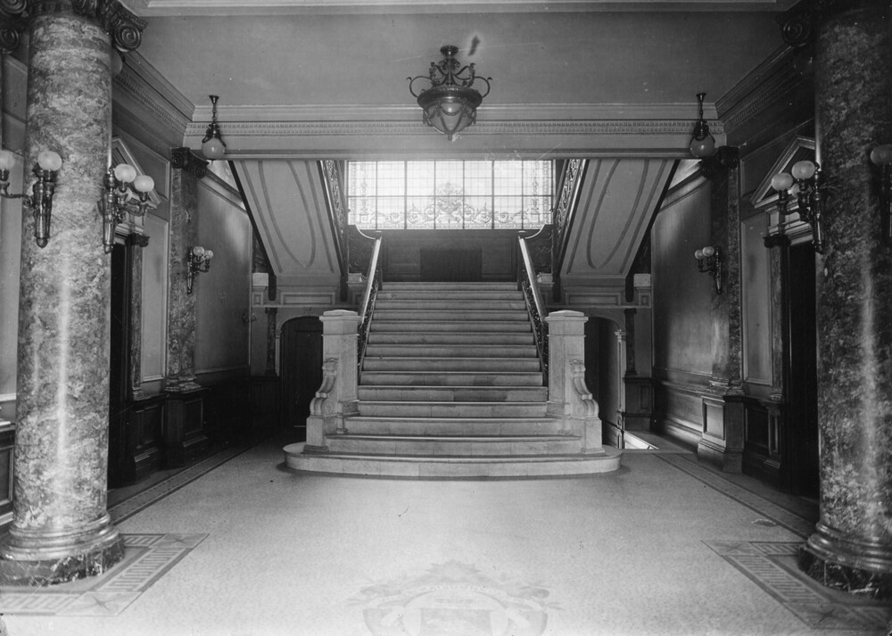 Black and white photograph of an entrance hall leading to a staircase. The hall has a Beaux-Arts look with marble pillars and white walls. Lights are attached to the pillars and suspended from the ceiling. On the floor, very pale tiled coat of arms can be seen.