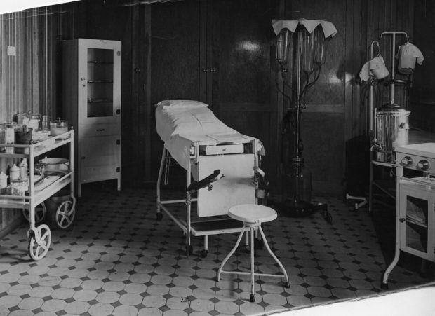 Black and white photograph of a room with lacquered wooden walls and tiled floors. The room is furnished with a hospital bed, cabinets, a wheeled table full of medical equipment, and several machines consisting of large glass and steel tanks.