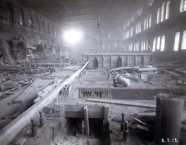 Wood beams lay on the floor, daylight shines through the numerous windows. Sand bags and debris lay across the unfinished floor.