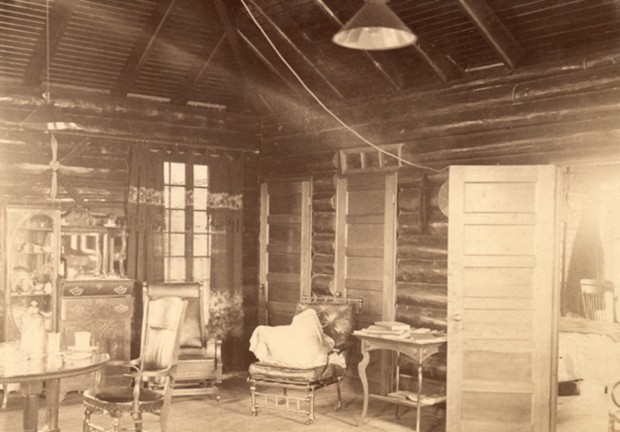 Blockhouse interior. Log cabin with ceiling lamp, one window, several chairs, a table and chest of drawers.