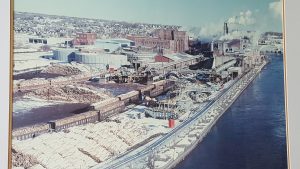 Photograph of the paper mill in full production. It's a very busy image, which includes a shot of the St. Mary's River, the train tracks running through the mill site, train cars full of logs, cut logs piled high by the river, the pulp tower and other buildings in full production, which is apparent by the smoke coming out of the smoke stacks.