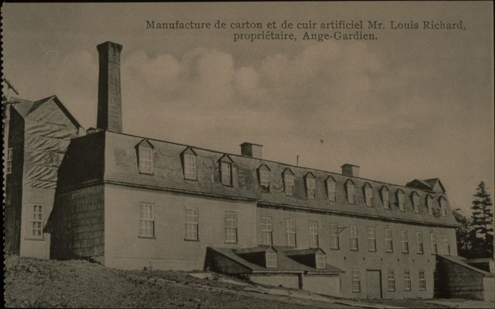 Black and white archival photo showing a three-quarter view of an industrial, three-floor building with multiple windows and a mansard roof. A tall chimney stands behind the building to the left. Two lower chimneys emerge from the centre of the factory.