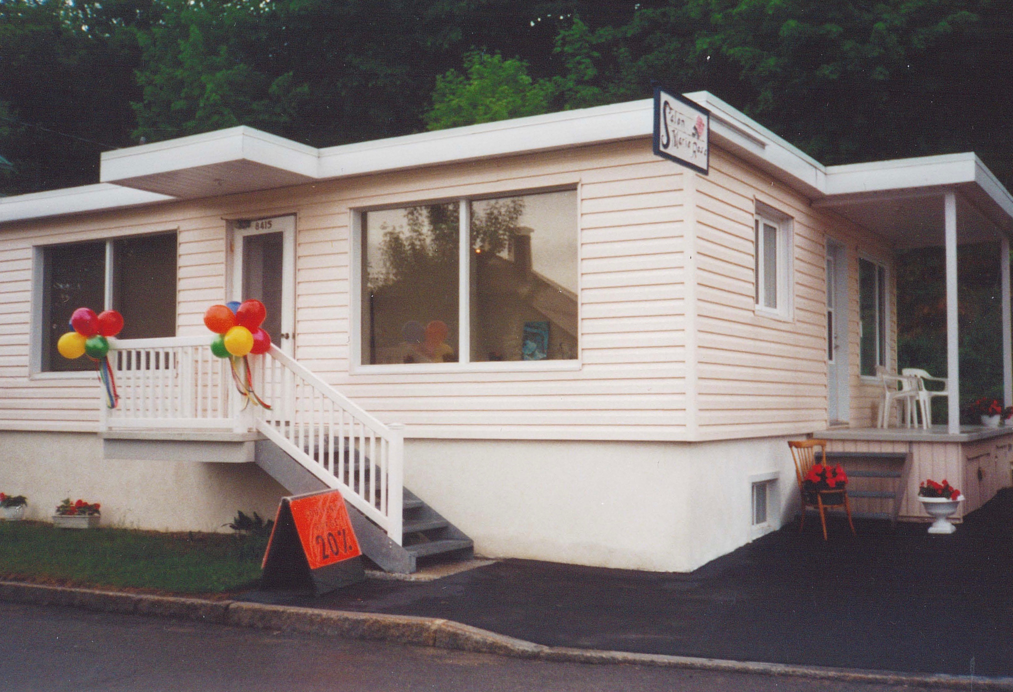 Colour photo of a single-storey, pale pink house. The three-quarter view shows two entrances, one at the side and another at the front. We can see a few white chairs on the little side porch, and a flowerpot containing red blooms at the foot of the side stairs. At the front of the building, bunches of red, yellow, green, and blue balloons are tied to the porch railing. An orange sign in front of the house advertises “Bras: 20% off.” A sign for Salon Marie-Rose hangs perpendicular to the façade, where it can be seen from the road.