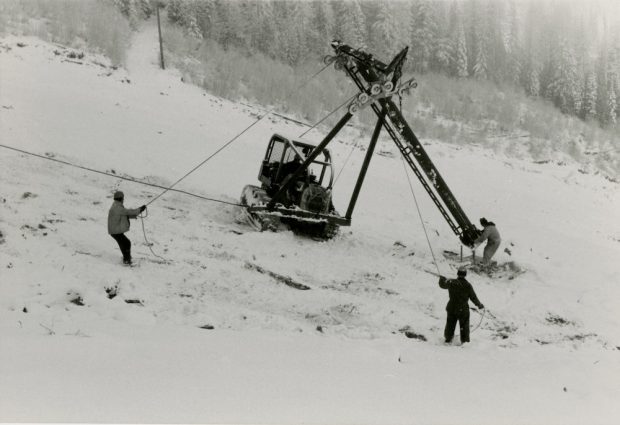 Black and white photograph of a crane and three men installing a tower on a ski slope.