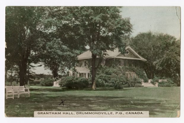 Black and white photograph of Grantham Hall, a large two-storey stone house covered in climbing plants with large stairs leading to the property’s gardens. The gardens consist of shrubs and mature trees.