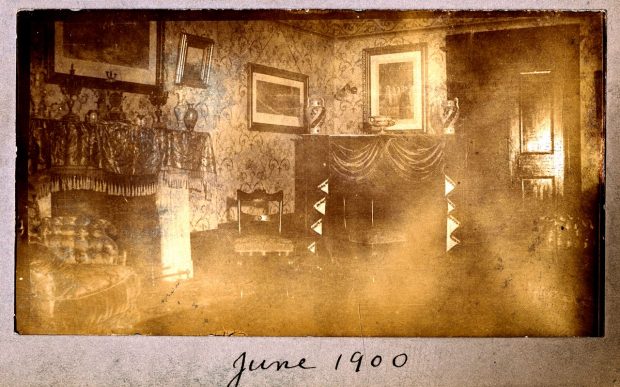 Black and white photograph of the living room of Grantham Hall decorated with framed pictures on the walls and draped furniture. A fireplace, chairs, and an armchair are visible.