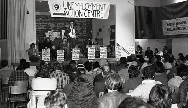 A man stands at a microphone to address an audience from a stage. Three men are seated on the stage . A banner above the stage reads “Unemployment Action Centre” and other signs are taped to the stage.