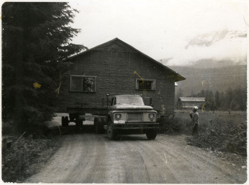 Black-and-white photograph showing a flatbed truck moving a house. The truck is on a road with a tree to the left, and a man standing on the right.