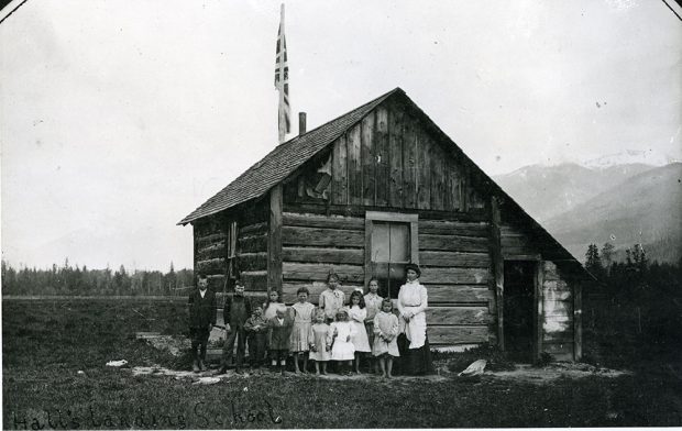 Black-and-white photograph. Twelve school children standing outside of a wooden schoolhouse with their teacher. The schoolhouse has a door on the right, a window on the front and side, and a flag pole at the back. The school sits on a small patch of snow, with trees in the background and faded mountains in the back right.