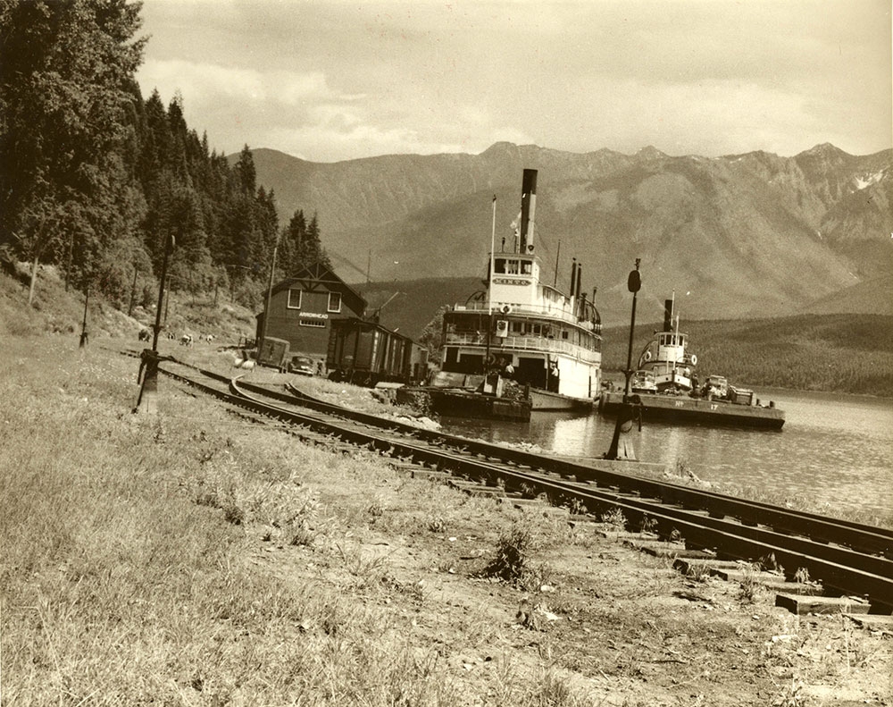 Two steamships are docked on the side of the river beside railway tracks in this black-and-white photograph. A small building sits at the end of the tracks by the boats. There are trees on the left and mountains in the background.