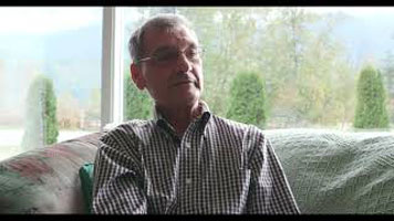 Brian Gawiuk - a white man with grey hair, glasses, and a plaid button down shirt. He is sitting in a living room on a couch, with clear windows behind him and a view of trees in the background.