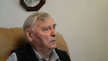 Harold Catherwood - an elderly white man with short grey hair. He is only shown from the shoulders up, and is facing the right. He is sitting on a light brown armchair in a room with black and white clock on a white wall.