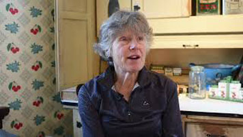 Nancy Martin – a white women with short grey hair, wearing a navy blue zip-up sweater. She is sitting in the kitchen area of the exhibit with artifacts and wallpaper behind her.