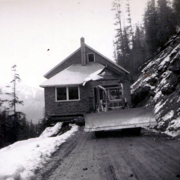 Black-and-white photograph of a small brick building being pulled by a snowplough across a dirt road. The building hangs slightly over the side of a cliff. Snow is on the ground, and trees are on either side of the road.