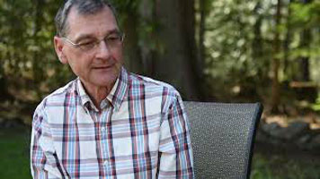 Brian Gawiuk - a white man with grey hair, glasses, and a plaid button down shirt. He is sitting outside on the edge of a grey chair with the forest behind him.