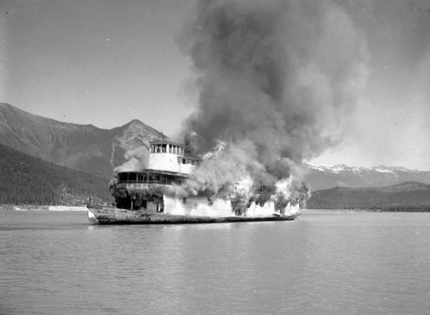 Black-and-white photograph of a steamship burning in the middle of a lake. Huge smoke clouds and flames are coming off the boat. Mountains are in the background.