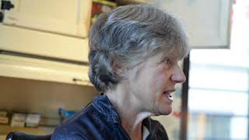 Nancy Martin – a white women with short grey hair, wearing a navy blue zip-up sweater. She is sitting facing right in the kitchen area of an exhibit with artifacts behind her.