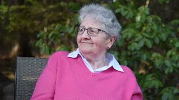 Marris - an elderly white woman, with white-grey hair, wearing black glasses, a white collared top, with a pink sweater over top.