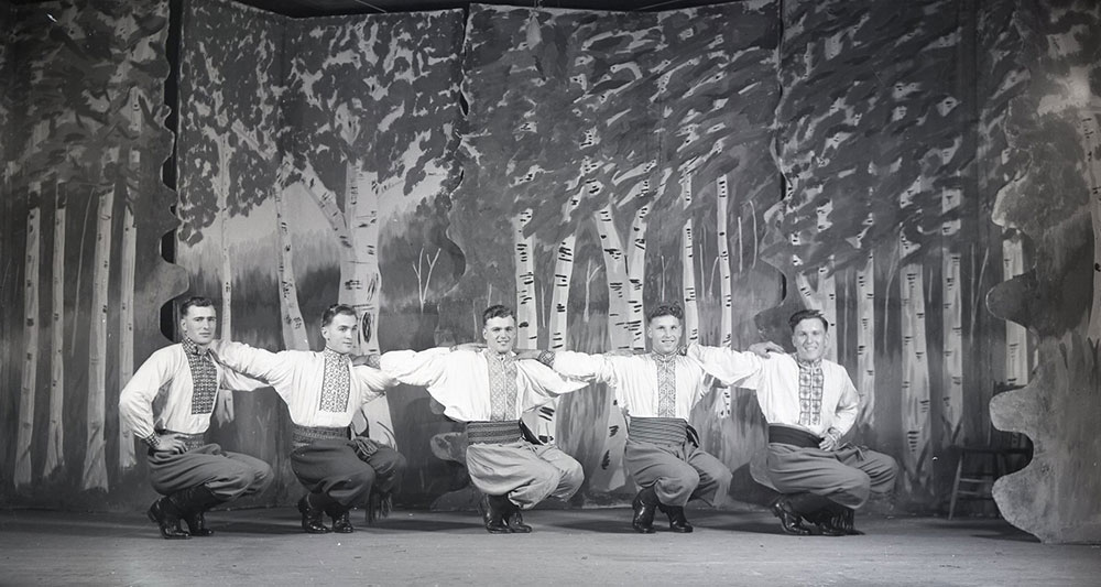 A black-and-white photograph of five dancers on a stage. The dancers are squatted down on their feet and have their arms on each others shoulders. The stage background is a mural of poplar trees.