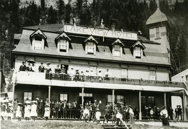 Black-and-white photograph of a two-storied hotel, with name Lakeview Hotel at the top. A crowd of people stand on and below the balcony. The people are in dress-attire - wearing suits, dresses, and hats.