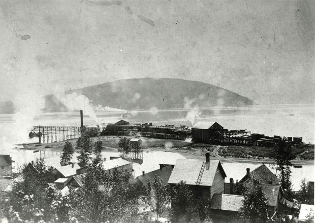 A black-and-white photograph of a town by the water. Trees and houses in the foreground. A lumber mill sits by the water, and there is smoke coming off the mill. A steamboat is in the distance on the water, with a mountain in the background.