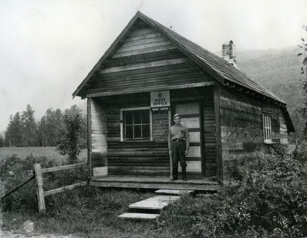 A black-and-white photograph of a man standing on the porch of a log building with a sign that reads Post Office Mount Cartier. The building is surrounded by greenery and trees, and has a wooden fence on the left.