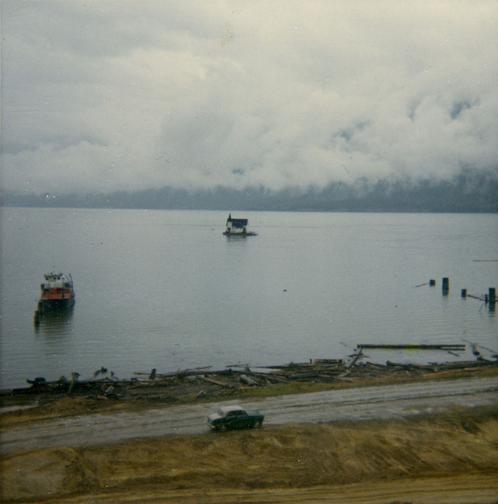 A colour photograph of a church surrounded by a body of water. A boat is on the left side of the photo in the water, and the tops of a dock are on the right. In front of the water a car drives on a road with wood piles on the left, and land on the right. Low clouds are in the background.